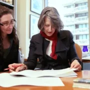 Two women sit at a desk in an office, reviewing documents together. One woman has gray hair and wears a red scarf, while the other has long dark hair and wears glasses.