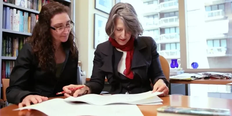 Two women sit at a desk in an office, reviewing documents together. One woman has gray hair and wears a red scarf, while the other has long dark hair and wears glasses.