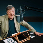 A person with gray hair and a beige jacket speaks into a microphone in a recording studio. A notebook, glasses, and a glass of water are on the desk.