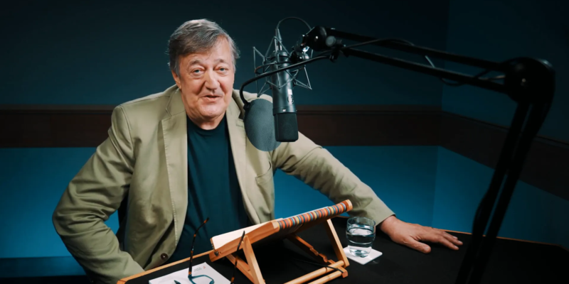 A person with gray hair and a beige jacket speaks into a microphone in a recording studio. A notebook, glasses, and a glass of water are on the desk.