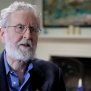 An older man with glasses and a beard speaks in an interview setting, with a fireplace and artwork visible in the background.