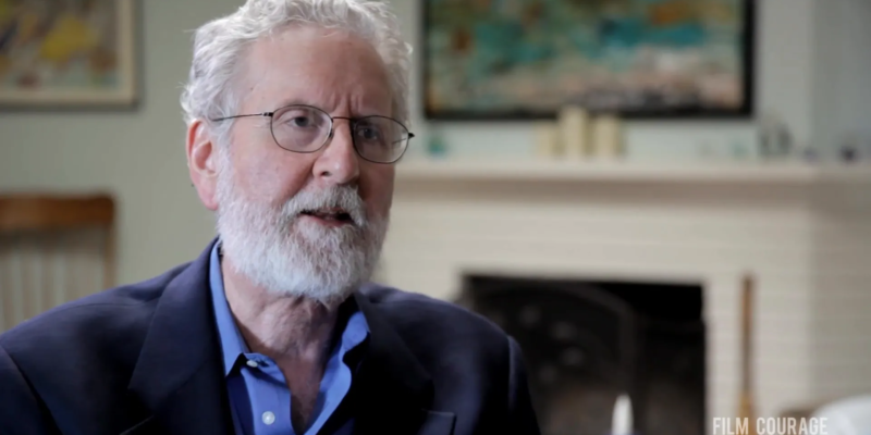 An older man with glasses and a beard speaks in an interview setting, with a fireplace and artwork visible in the background.