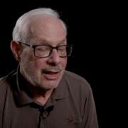 An older man with glasses and a short beard speaks against a dark background. He's wearing a brown polo shirt.