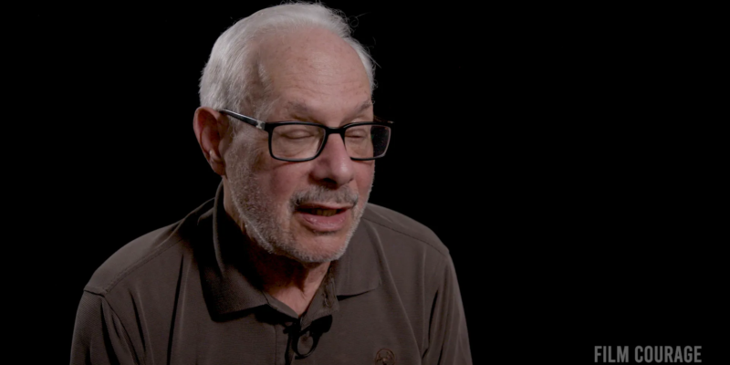 An older man with glasses and a short beard speaks against a dark background. He's wearing a brown polo shirt.