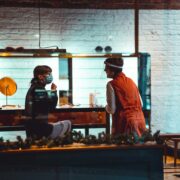 Two people engage in a conversation at a table inside a brightly lit room, one wearing a red outfit and the other sporting a mask reminiscent of writing's mysteries, with golden decor providing captivating insights into the scene.