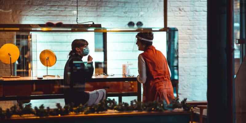 Two people engage in a conversation at a table inside a brightly lit room, one wearing a red outfit and the other sporting a mask reminiscent of writing's mysteries, with golden decor providing captivating insights into the scene.