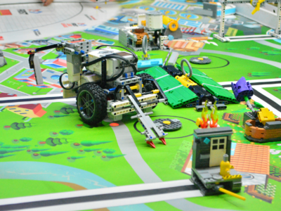 Robotics competition setting with colorful toy vehicles and structures on a map-like board. A central robot features wheels and a protruding apparatus. Another robot nearby has green and yellow pieces. Various small models depict scenes of a city environment with roads and buildings.