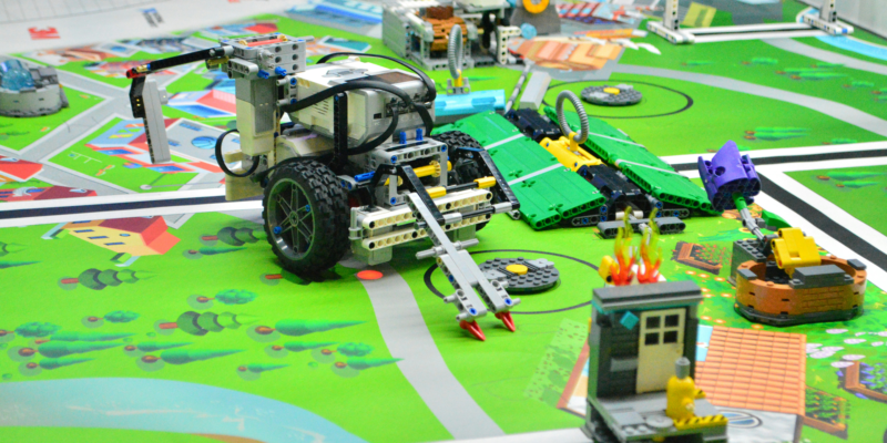 Robotics competition setting with colorful toy vehicles and structures on a map-like board. A central robot features wheels and a protruding apparatus. Another robot nearby has green and yellow pieces. Various small models depict scenes of a city environment with roads and buildings.