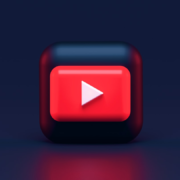 A 3D icon featuring a red square with a white play button in the center, resembling the YouTube logo, set against a dark blue background. The icon has a glossy finish, casting a subtle reflection on the surface below.