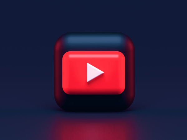 A 3D icon featuring a red square with a white play button in the center, resembling the YouTube logo, set against a dark blue background. The icon has a glossy finish, casting a subtle reflection on the surface below.