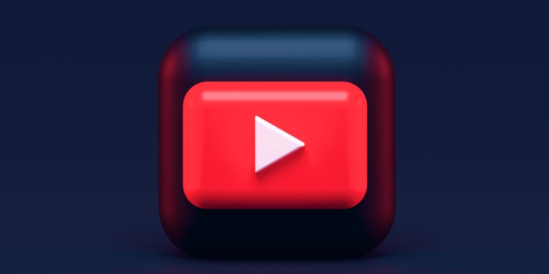 A 3D icon featuring a red square with a white play button in the center, resembling the YouTube logo, set against a dark blue background. The icon has a glossy finish, casting a subtle reflection on the surface below.