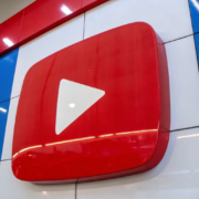 A large 3D YouTube logo with a red play button is displayed on a wall with a checkered blue, white, and red pattern. The logo stands out prominently, and overhead fluorescent lights are reflected on the shiny surface of the wall.