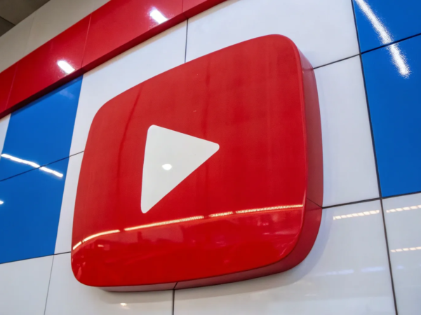 A large 3D YouTube logo with a red play button is displayed on a wall with a checkered blue, white, and red pattern. The logo stands out prominently, and overhead fluorescent lights are reflected on the shiny surface of the wall.