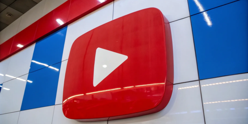 A large 3D YouTube logo with a red play button is displayed on a wall with a checkered blue, white, and red pattern. The logo stands out prominently, and overhead fluorescent lights are reflected on the shiny surface of the wall.